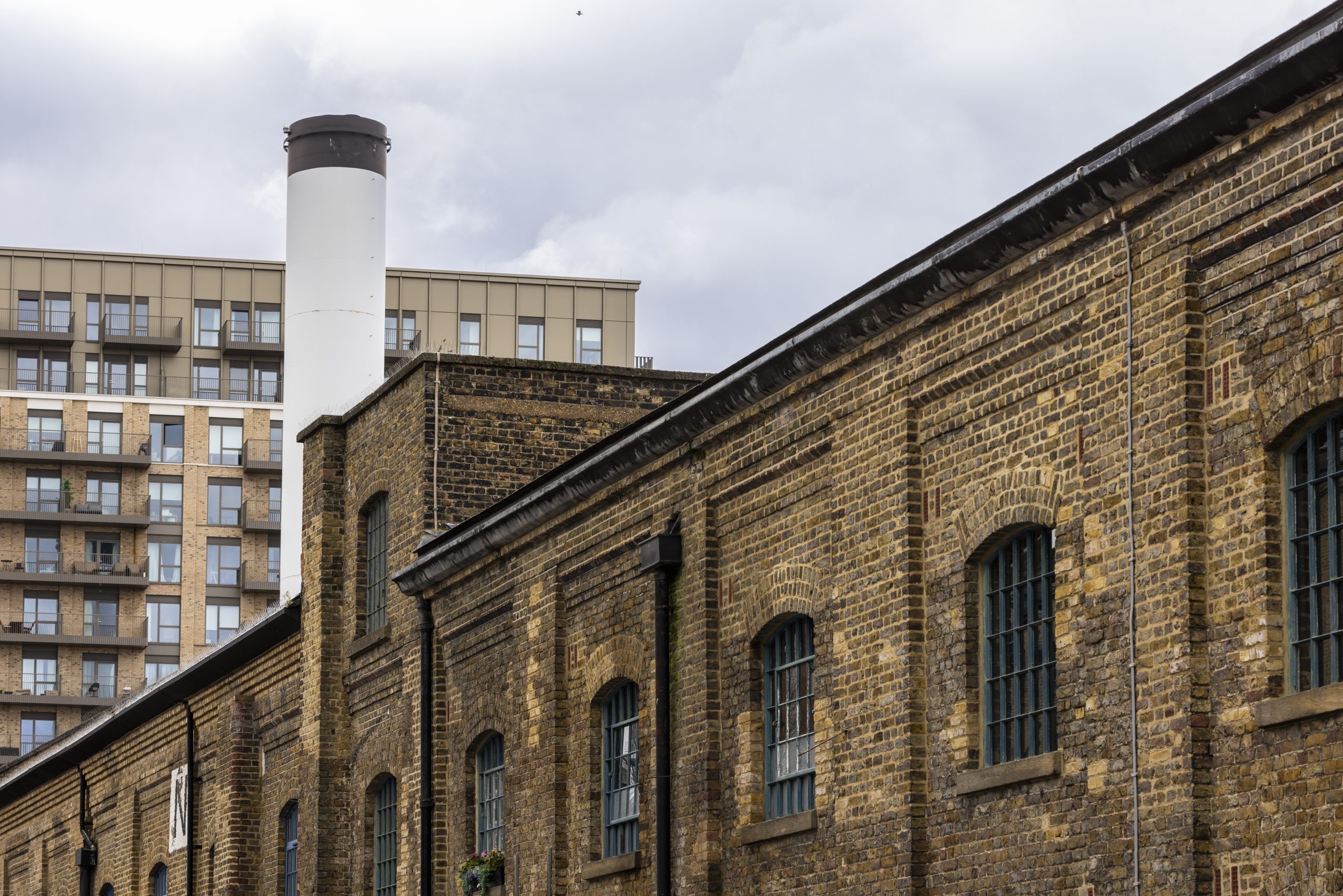 Old brick industrial building with arched windows in foreground, modern apartment block and a tall white chimney rising in background under a cloudy sky.