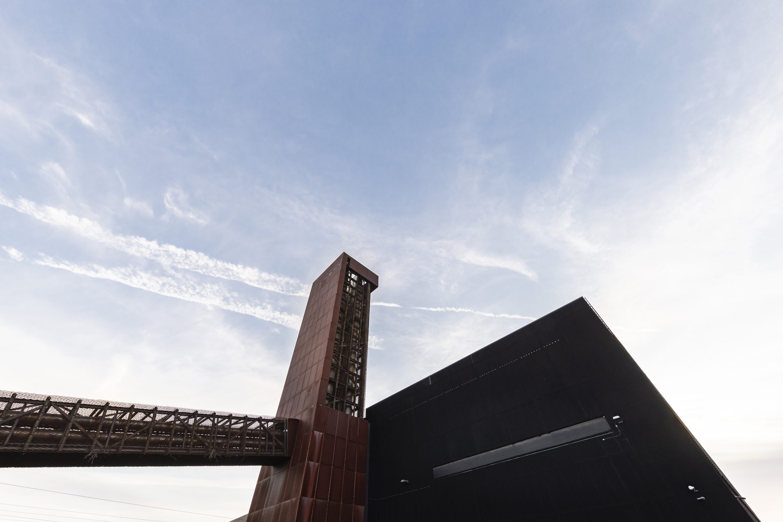A modern industrial building with a tall, rust-coloured tower and metal bridge under a clear blue sky.