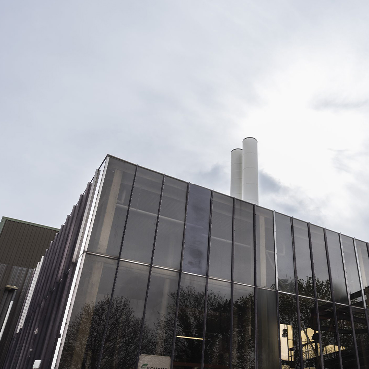 Industrial building with large glass panels and two tall white chimneys under a cloudy sky. Reflective surfaces create a stark, modern atmosphere.