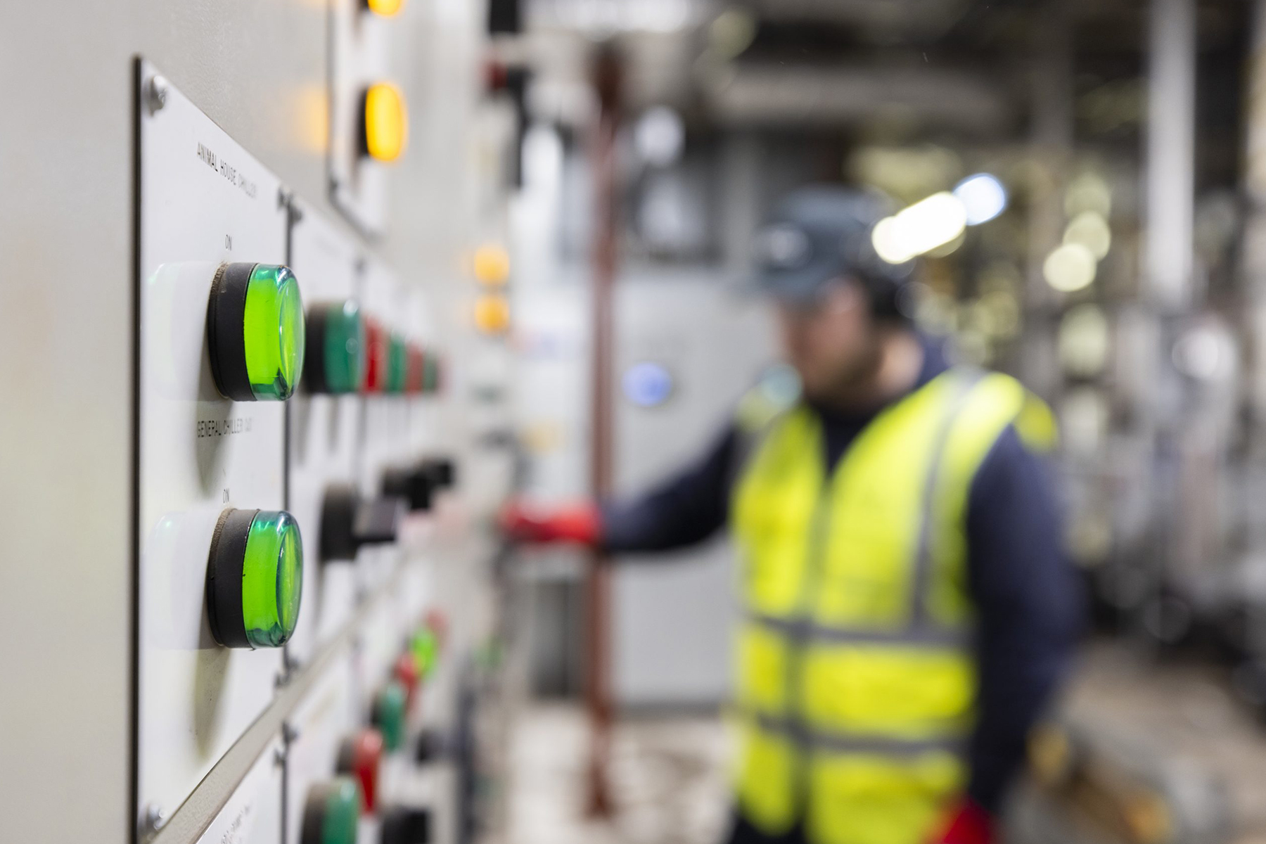 Close-up of a control panel with green and orange buttons. In the blurred background, a worker in a yellow vest and helmet operates machinery. Industrial setting.