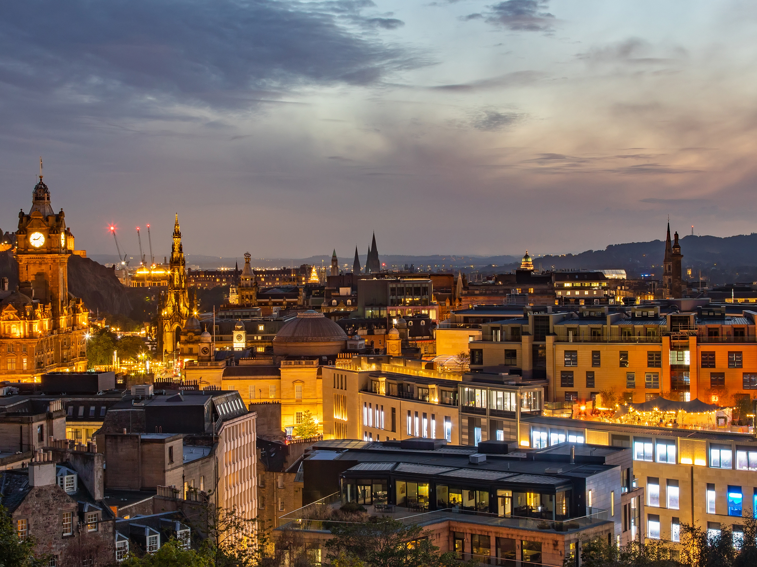 A cityscape of Edinburgh at dusk, with softly lit buildings, including a castle on a hill. The sky is a blend of oranges and purples.