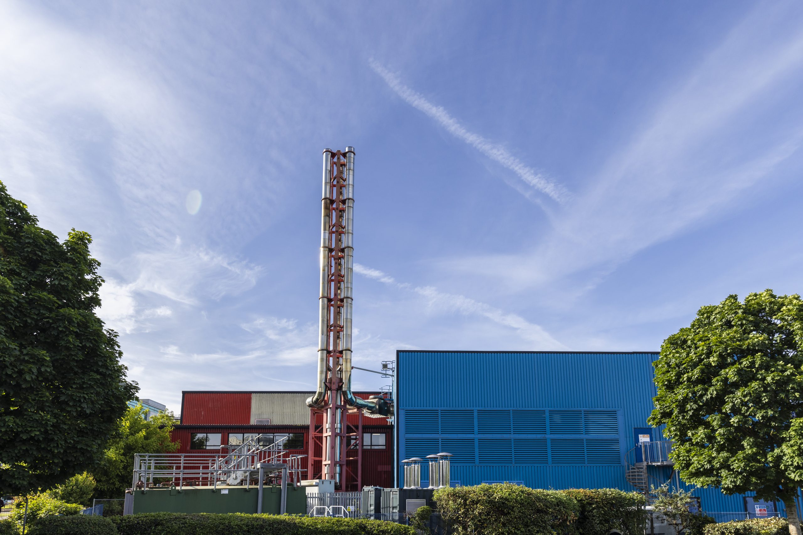 Industrial building with a tall, striped chimney against a clear blue sky. The structure features blue and red sections, flanked by green trees.