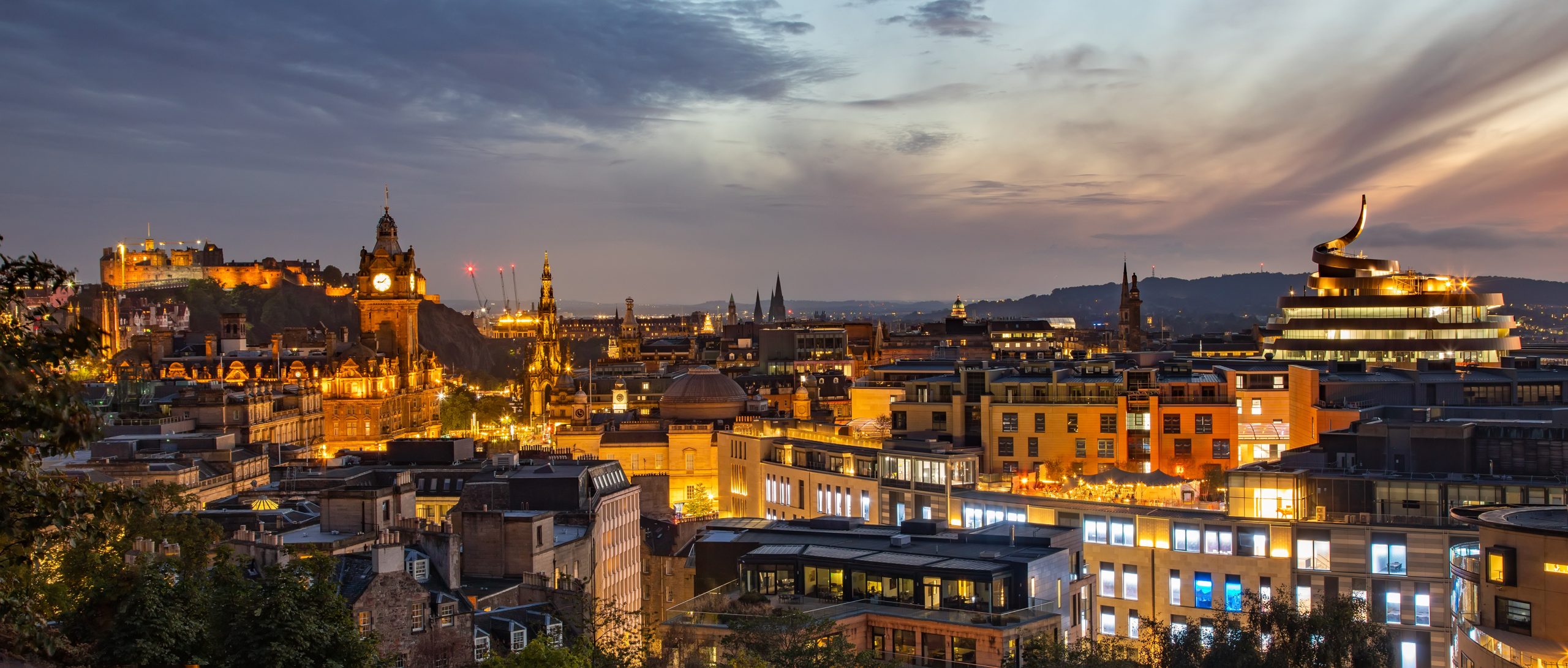 A cityscape of Edinburgh at dusk, with softly lit buildings, including a castle on a hill. The sky is a blend of oranges and purples.
