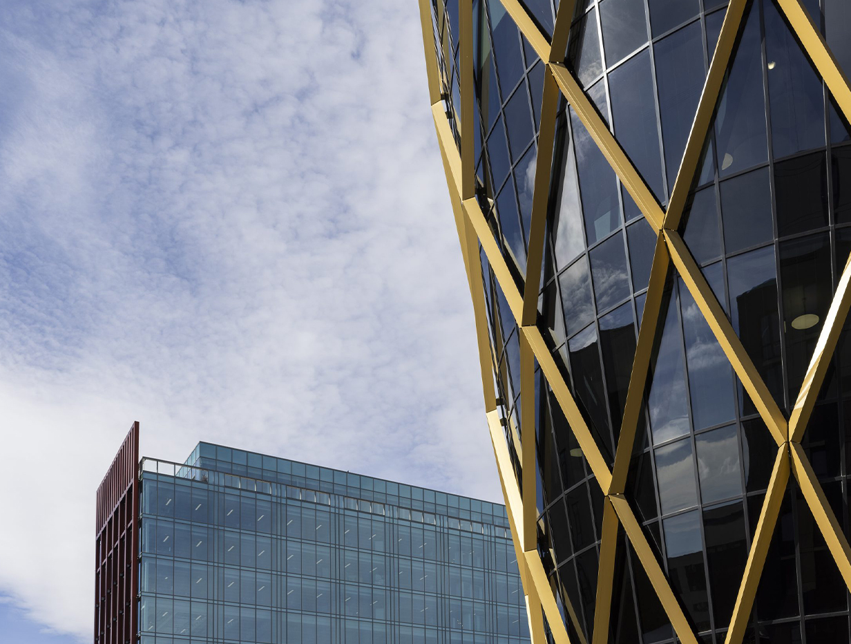 Modern buildings under a partly cloudy sky. One structure features gold lattice framing with glass panels, while another has blue-tinted glass and a red accent.